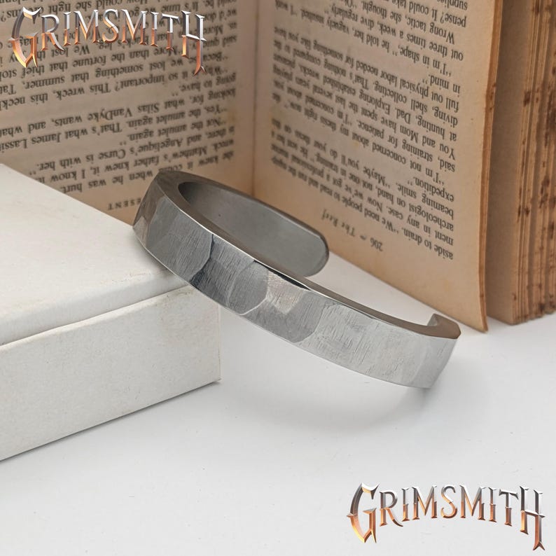 May include: A silver-toned cuff bracelet with a hammered texture. The open-ended design features a flat, wide band with a slightly curved shape. The bracelet is displayed on a white surface with a book in the background. The Grimsmith logo is visible.
