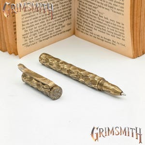 Peut inclure: Un ensemble de stylo et de clip de stylo avec un motif textur&eacute; unique dans des tons or et marron. Le stylo a une pointe m&eacute;tallique &eacute;l&eacute;gante et le clip pr&eacute;sente un design articul&eacute;. Le mot "GRIMSMITH" est affich&eacute; en bas &agrave; droite.