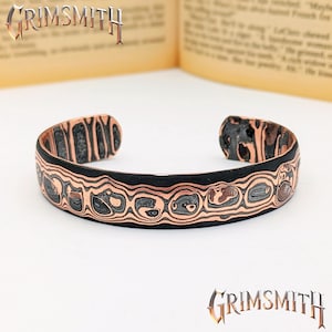 May include: A copper cuff bracelet with a black border and intricate patterned design. The bracelet features a series of circular and oval shapes with a textured appearance. The word "GRIMSMITH" is displayed in a stylized font.