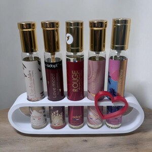 Peut inclure: Présentation de cinq flacons de parfum avec des bouchons dorés, chacun avec une étiquette et un design différents, disposés dans un support blanc. Les bouteilles sont étiquetées "L'or des sables", "Love mood", "ROUGE", "COTTON" et "PARADISE". Une décoration rouge en forme de cœur est devant.