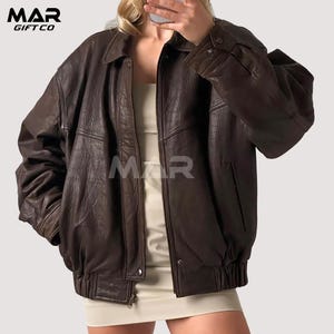 Può includere: Giacca in pelle marrone con zip, colletto e polsini con bottoni. La giacca ha un design in stile bomber con orlo a coste e tasche. Il logo MAR GIFT CO è visibile in alto a sinistra e il logo MAR al centro.