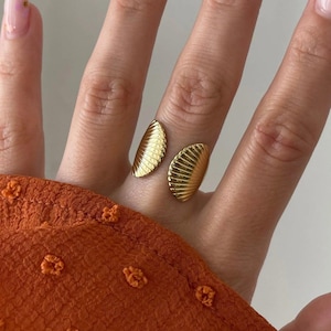 Puede incluir: Un anillo abierto de tono dorado que presenta dos extremos simétricos en forma de abanico con finos detalles acanalados verticales. El anillo se lleva en el dedo medio, posicionado contra una manga de tela naranja quemado con puntos.