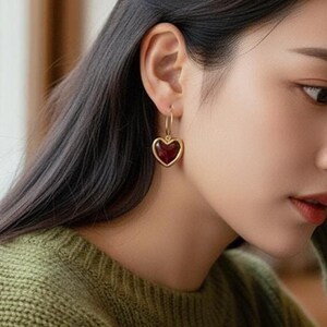 以下が含まれることがあります： 深い赤色の中心を持つ、金色のハート型イヤリング。イヤリングは、小さなフープとハートのペンダントが特徴のクラシックなデザインです。イヤリングは、グリーンのセーターを着た人に着用されています。