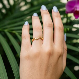 Può includere: Un anello color oro con un design ondulato è indossato su un dito. La mano è posta su uno sfondo di fogliame verde e orchidee viola. Il design dell'anello assomiglia a un'onda continua.