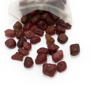 May include: A transparent plastic bag, slightly open, containing a collection of small, irregularly shaped, reddish-brown gemstones. The gemstones are scattered, with varying shades of brown and red. The background is plain white.