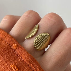 Puede incluir: Un anillo dorado con diseño de concha. El anillo presenta dos elementos de concha, uno más pequeño y otro más grande, con texturas acanaladas detalladas. El anillo se lleva en un dedo, con tela naranja en el fondo.