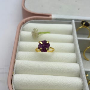 Pode incluir: Um anel dourado com uma gema bruta de cor magenta profunda. O anel tem uma configuração simples de quatro garras. O anel é exibido em uma caixa de joias com outros anéis e uma pequena flor branca.
