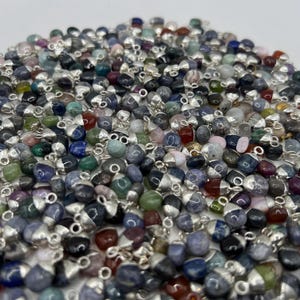 May include: A pile of small gemstone pendants with silver-coloured caps. The pendants are in various shapes and sizes, with a mix of colours including blue, green, pink, and red. The pendants are scattered across a white surface.