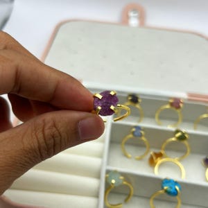 May include: A gold-coloured ring featuring a raw, uncut, purple gemstone held by four prongs. The ring is adjustable. Other rings with various coloured gemstones are visible in the background.