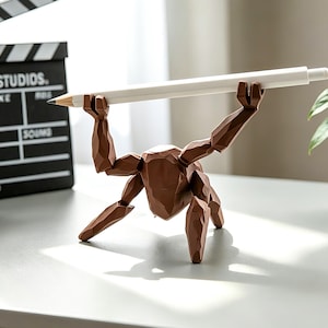 May include: A brown wooden figurine holds a white pencil overhead. The figurine has articulated limbs and stands on a gray surface. A white alarm clock and a film clapperboard are in the background.