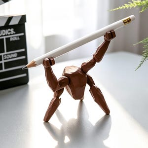 May include: A brown wooden artist's mannequin holding a white pencil with a gold ferrule. The mannequin is posed as if lifting the pencil. A black film clapperboard is in the background.