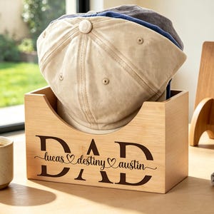 May include: A light-coloured wooden storage box featuring the word DAD engraved in large letters, with the names lucas, destiny, and austin separated by heart symbols. The box holds a stack of baseball caps in beige, blue, and dark grey colours.