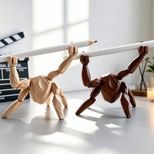 May include: Two wooden artist mannequins, one light and one dark, each holding a large white pencil. The figures are posed as if lifting the pencils. A film clapperboard and a clock are in the background, suggesting a creative or artistic theme.