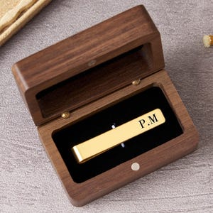 May include: A gold-toned metal tie bar with the engraved letters P.M. resting inside a rectangular dark brown wooden box with a black velvet interior lining. The box features a hinged lid and a small circular magnetic closure on the front edge.