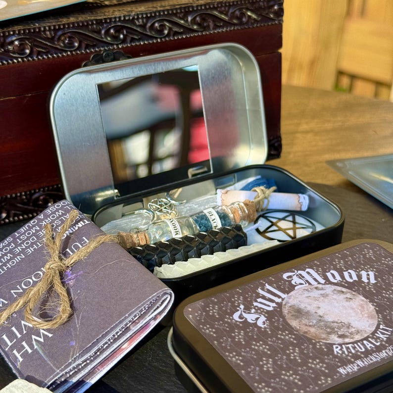 May include: An open, rectangular tin with a mirror inside, containing small glass bottles, black candles, and a white pentagram. A small booklet tied with twine and a brown tin labeled "Full Moon Ritual Kit" are also present.