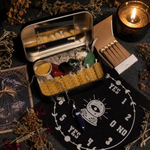 May include: A black metal tin containing yellow honeycomb-textured wax candles, assorted polished gemstones, and a small metal cup of yellow powder. A blue crystal pendulum on a silver chain rests on a black cloth with white text and symbols.