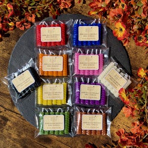 May include: A collection of hand-rolled beeswax spell candles in various colors, including red, blue, pink, orange, yellow, purple, green, brown, black, and white. Each set is packaged in a clear plastic bag with a label that reads "White Witch".