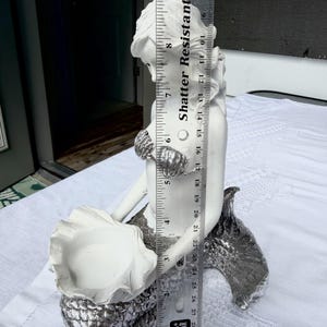 Op de afbeelding: Een wit en zilverkleurig zeemeerminbeeldje dat een schelpvormige kom vasthoudt. Een doorzichtige plastic liniaal is verticaal tegen het beeldje geplaatst en toont een hoogte van ongeveer 29 cm van de basis tot de bovenkant van het hoofd.