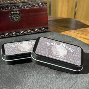 May include: Two black rectangular tins with white trim, labeled "Full Moon Ritual Kit." The lids feature a moon graphic and text. A dark wooden box with ornate metalwork is in the background. The tins are on a dark gray surface.