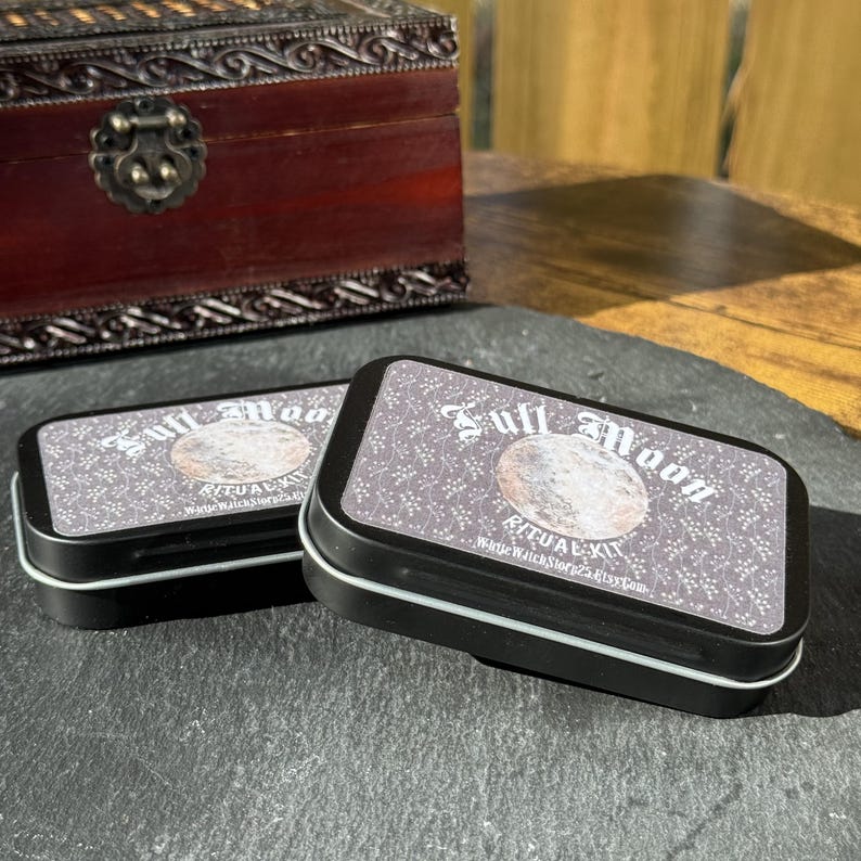May include: Two black rectangular tins with white trim, labeled "Full Moon Ritual Kit." The lids feature a moon graphic and text. A dark wooden box with ornate metalwork is in the background. The tins are on a dark gray surface.