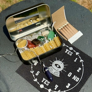 May include: A black metal tin filled with small candles, crystals, and a small container of sand. A pendulum necklace and a matchbook are also present. A black cloth with a white design of a clock face with the words "YES" and "NO" is in the foreground.