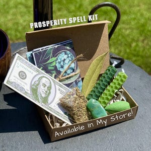 Może przedstawiać: Brązowe kartonowe pudełko z napisem PROSPERITY SPELL KIT oraz Available In My Store! zawierające replikę banknotu 100-dolarowego, woreczek suszonych zi&oacute;ł, zielony liść, dwie sześciokątne zielone świece woskowe oraz gładki zielony kamień.