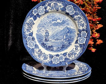 May include: A stack of blue and white ceramic plates. The plates feature a scenic landscape with a ship, mountains, and water. The rims are decorated with floral patterns. The plates are displayed on a clear stand.