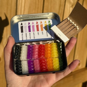 Puede incluir: Una caja metálica que contiene un juego de velas hexagonales de colores. Las velas están dispuestas en un espectro de arcoíris, con colores como negro, blanco, morado, rosa, rojo, naranja, amarillo y verde. Una pequeña caja de cerillas está abierta junto a la caja. La caja tiene el texto "Protection, Cleansing, Intuition, Self-Love, Love, Luck, Success, Prosperity."
