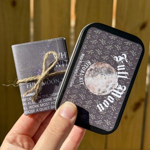 May include: A Full Moon Ritual Kit, featuring a small, dark gray booklet tied with twine and a rectangular tin. The tin has a starry background, a full moon graphic, and the words "Full Moon" in white script. The kit is designed for spiritual practices.