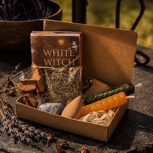 May include: A cardboard box containing a book titled White Witch, a clear plastic bag of dried herbs, a clear plastic bag of wood pieces, a blue stone, a tan cone-shaped incense, and two textured beeswax candles in green and orange colors.
