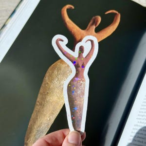 May include: A sticker featuring a stylized figure with arms raised, adorned with colorful holographic accents. The figure is brown with a white border, set against a dark background. The sticker's design is reminiscent of ancient art.