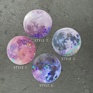 May include: Four round stickers with moon designs. Style 1 is pink and purple with stars. Style 2 is purple with glitter. Style 3 is blue and gray with green glitter. Style 4 is purple and blue with stars.