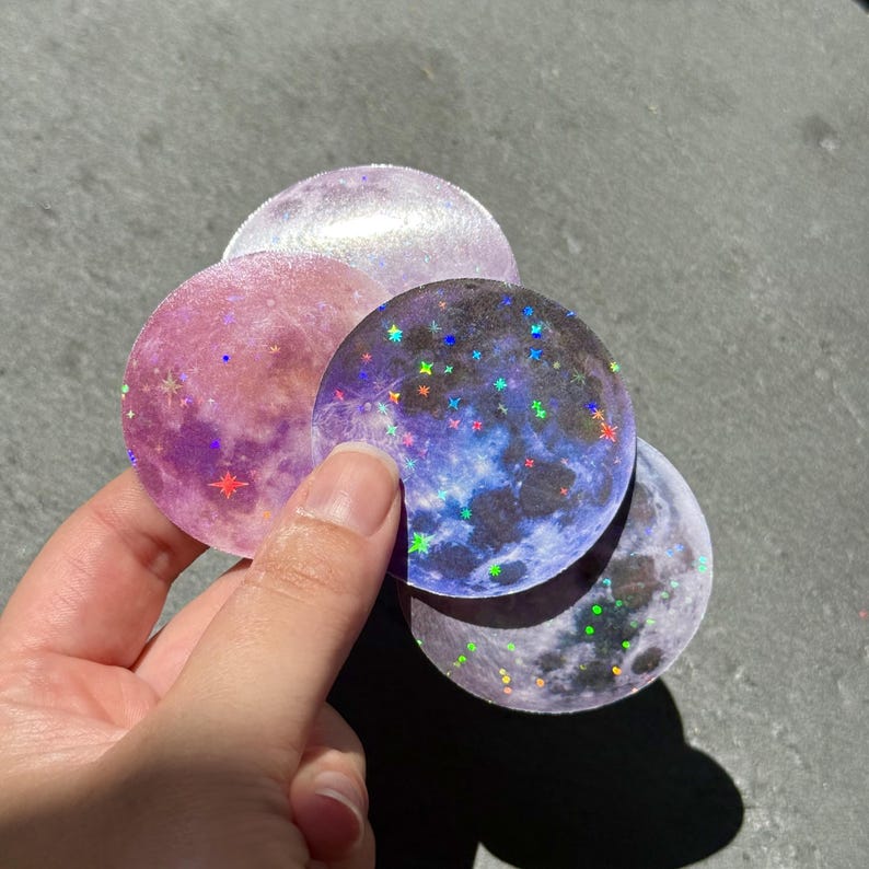May include: A hand holds a collection of round stickers with a celestial theme. The stickers feature holographic moon designs in shades of pink, purple, and blue, with scattered star accents. The stickers have a shiny, iridescent finish.