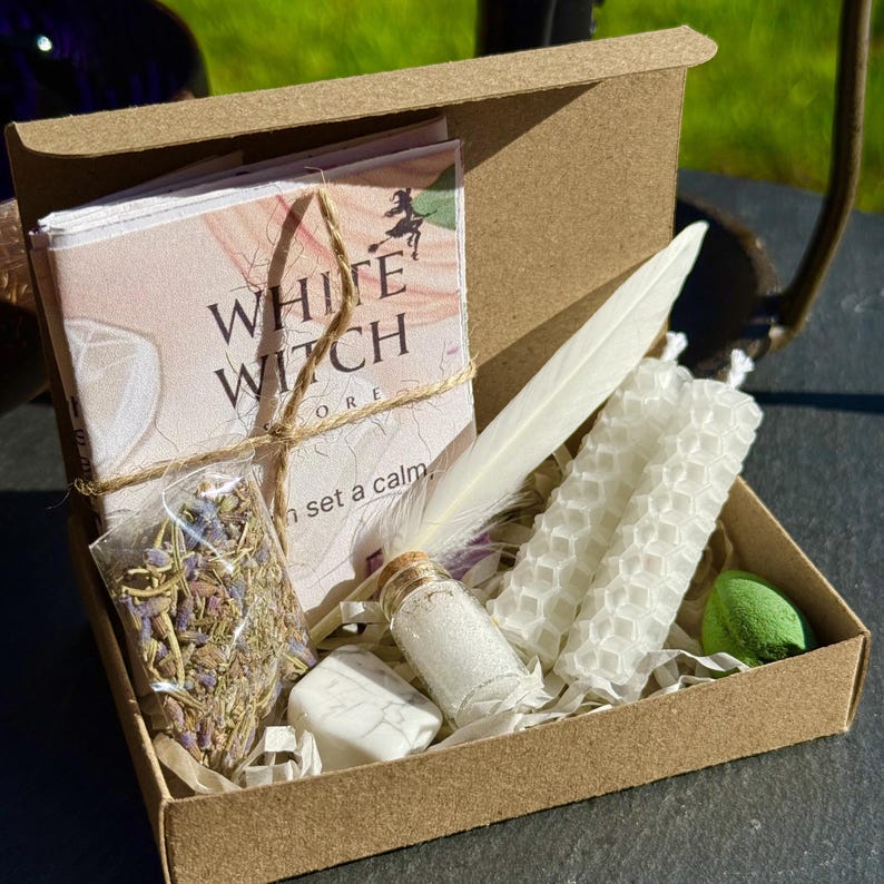 Może przedstawiać: Brązowe kartonowe pudełko zawierające kartę White Witch Store, mały plastikowy woreczek z suszoną lawendą, białe pi&oacute;ro, dwie białe świece o strukturze plastra miodu, małą szklaną fiolkę z białym proszkiem oraz biały kamień z szarymi żyłkami.
