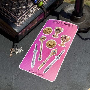 May include: A sticker sheet with tarot symbols, including swords, chalices, and wands, in gold and silver. The sheet has a pink and purple gradient background. A decorative journal and charms are also visible.