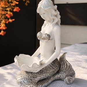 Op de afbeelding: Een wit en zilverkleurig zeemeerminbeeldje met een matwit bovenlichaam, een metallic zilveren geschubde staart en een schelpvormige top. De zeemeermin houdt een grote, open witte schelpvormige kom in haar handen als decoratieve schaal.