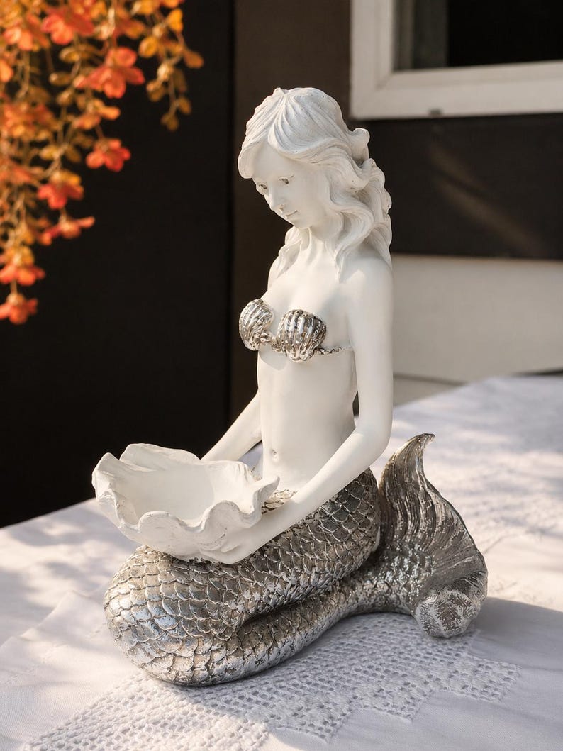 Op de afbeelding: Een wit en zilverkleurig zeemeerminbeeldje met een matwit bovenlichaam, een metallic zilveren geschubde staart en een schelpvormige top. De zeemeermin houdt een grote, open witte schelpvormige kom in haar handen als decoratieve schaal.