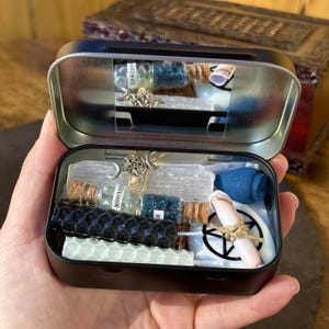 May include: Open black metal tin filled with various spiritual items. Includes small glass bottles with corks, blue crystals, white candles, a blue incense holder, and a rolled scroll tied with twine. A silver pentacle charm is visible.