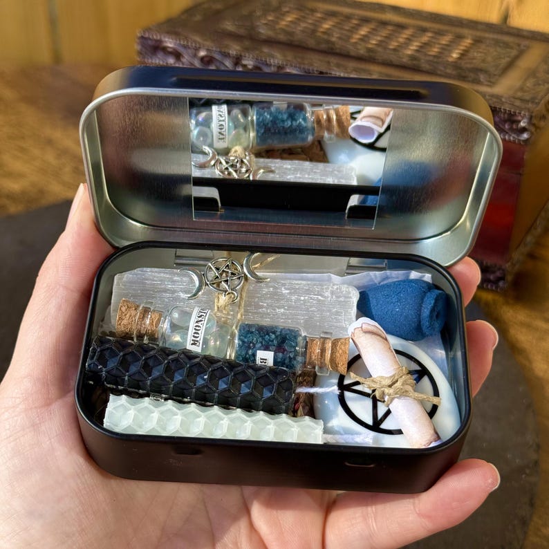May include: Open black metal tin filled with various spiritual items. Includes small glass bottles with corks, blue crystals, white candles, a blue incense holder, and a rolled scroll tied with twine. A silver pentacle charm is visible.