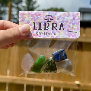 May include: A clear plastic bag containing a Libra crystal set. The set includes five polished stones in various colors: pink, green, blue, and gray. The bag has a decorative header with the word "LIBRA" and a scale symbol.