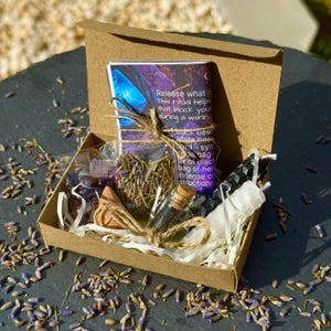May include: A small, open cardboard box filled with various items, including a small glass bottle with a cork, a black candle, and a bundle of dried lavender. The box also contains a card with text and a small cone-shaped object. Dried lavender is scattered around the box.