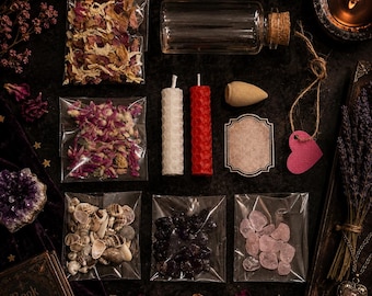 May include: A collection of ritual supplies including dried flower petals in clear bags, a glass vial with a cork, two honeycomb-textured candles in white and red, an incense cone, a blank label, a pink heart tag, seashells, and crystals.