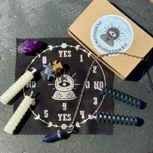 May include: A pendulum divination kit with a black mat featuring a crystal ball graphic and the words "YES" and "NO". Includes a blue-speckled pendulum, two black hexagonal candles, two cream-colored candles, a purple crystal, and a small glass bottle. A cardboard box is also present.