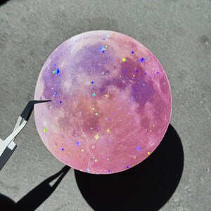 May include: A round, pink and purple moon-shaped sticker with iridescent star-shaped glitter. The sticker has a gradient effect, transitioning from pink to purple. A pair of tweezers is holding the sticker.