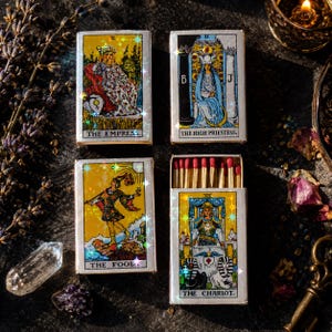 May include: Four rectangular matchboxes featuring tarot card illustrations titled The Empress, The High Priestess, The Fool, and The Chariot. The boxes have a holographic finish and one box is open, revealing wooden matches with red tips.