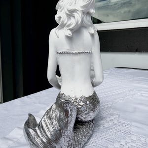 Op de afbeelding: Een wit zeemeerminbeeldje met een metallic zilveren staart met gestructureerde schubben. Het figuur heeft lang, golvend wit haar en is van achteren afgebeeld, met een glad bovenlichaam en een gedetailleerde, glinsterende zilveren staartvin.