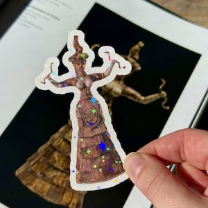May include: A holographic sticker depicting the Snake Goddess of Knossos. The sticker shows a woman with outstretched arms, holding snakes, and wearing a tiered skirt. The sticker has a white border and is held by a hand.
