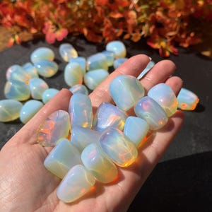 May include: A collection of smooth, tumbled opalite stones with a translucent, milky white appearance and iridescent blue and orange light reflections. The stones are rounded, polished, and exhibit a glass-like texture with internal glow.