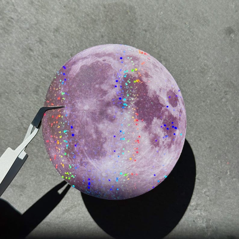 May include: A purple, full moon-shaped object with glitter accents in various colors. The moon has craters and other surface details. A pair of tweezers is positioned near the moon's surface, casting a shadow.