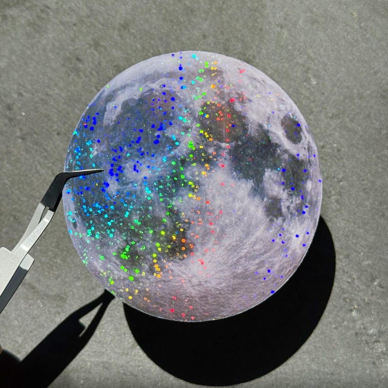 May include: A round, moon-shaped sticker with a detailed surface, speckled with colorful glitter. The sticker is held by tweezers, casting a shadow on a gray surface. The glitter includes blue, green, yellow, and red hues.